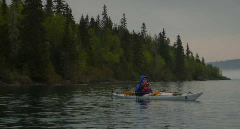 a student sits in a kayak on a calm lake lined with trees